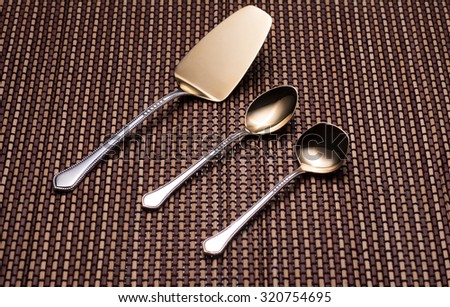 Shining silver kitchen utensils with golden spraying at the end. Shovel cake, teaspoon and spoon for sugar on the pattern table mat.