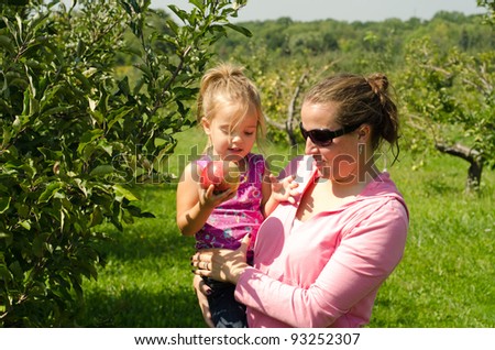 Half body portrait of happy mother holding preschool daughter in orchard, child with ripe apple.