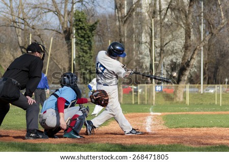 ZAGREB, CROATIA - MARCH 28, 2015: Baseball match Baseball Club Zagreb in white jersey and Baseball Club Nada in blue jersey. Baseball batter, catcher and plate umpire. Ball flying through the air