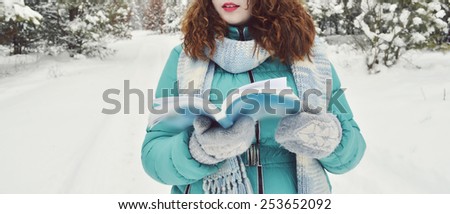 young,beautiful,curly,dark-haired girl dressed in a turquoise jacket,bodily woolly hat, and a light blue scarf,reading a book in the Park,drinking tea,coffee,cocoa