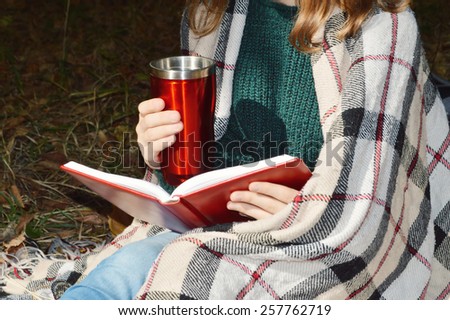 a young girl wrapped in a warm plaid, reading a book in the Park.hot drinks, warming, herbal tea.Autumn concept .	\
 spring,walk,Park,nature