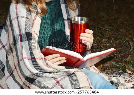 a young girl wrapped in a warm plaid, reading a book in the Park.hot drinks, warming, herbal tea.Autumn concept .	\
 spring,walk,Park,nature