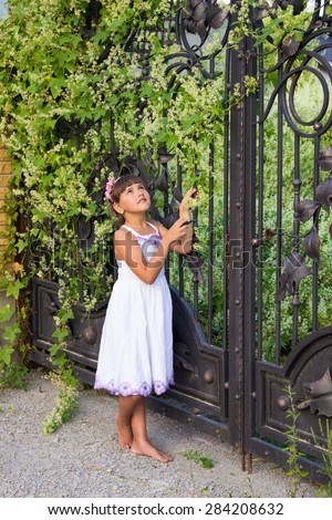 little girl, eight years old, brunette with blue-green eyes in a white dress standing near a large wrought-iron gates