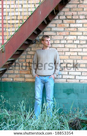 Young stylish man with blonde hair stay under stairs near brick wall.