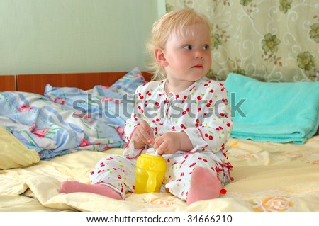 Pretty little girl in pyjamas (pajamas) with feeding bottle sit on the unmade bed.