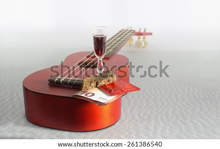 Close view of a small guitar ukulele, a bread, ten euro banknote, a small glass with a drink and a condom at seaside Ã¢?? six simple things for a better life.