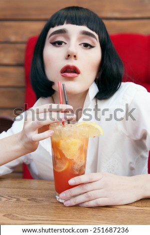 Cute woman in a black wig and the white shirt. Closeup portrait of a young cute girl over red background. Perfect bright makeup. Red lips. Pulp Fiction. With cocktail.