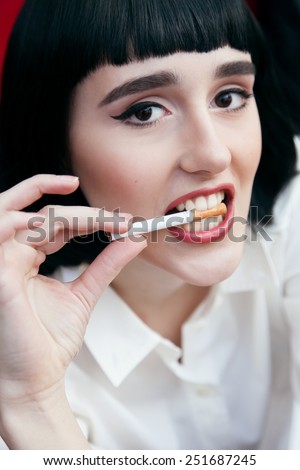 Beautiful cute woman in a black wig and the white shirt. Closeup portrait of a young cute girl over red background. Perfect bright makeup. Red lips. Pulp Fiction. With cigarette.