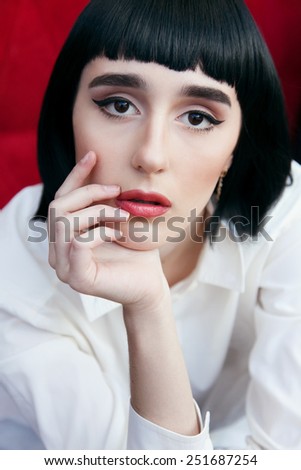 Beautiful woman in a black wig and the white shirt. Closeup portrait of a young cute girl over red background. Perfect bright makeup. Red lips. Pulp Fiction.