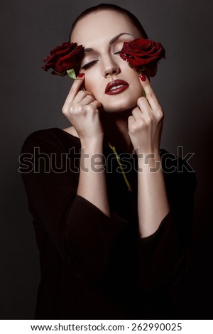 Beautiful brunette with red lips holding red roses by her face. Black dress. Studio shot. Black background