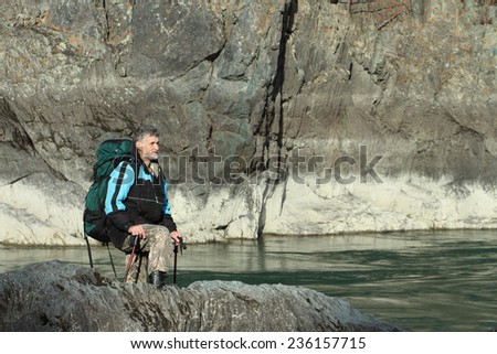 The traveler  with a backpack and tracking sticks going on the rocky bank of the mountain river