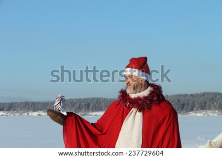 The man in Santa Claus\'s suit the bag holding in hand with a gift against the blue sky and a snow field with trees