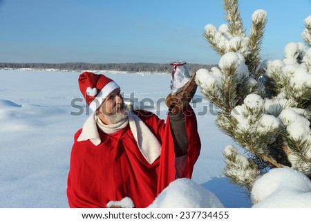 The man in Santa Claus's suit the bag holding in hand with a gift against the blue sky and a snow-covered pine