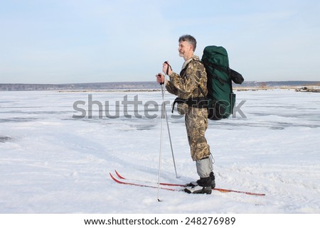 The man the traveler with a backpack standing on skis on the snow river bank