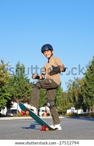 Smiling teenage boy with his thumbs up standing on a skateboard on a sunny day with blue sky and trees in the background.