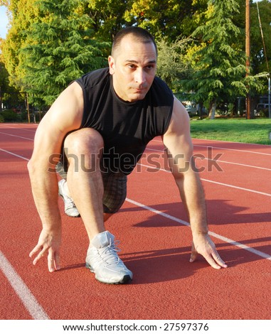 Male athlete getting ready to run on an athletic track with trees in the background on a sunny day.