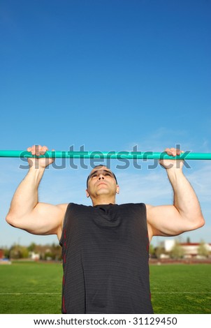 Strong man doing pull ups on a bar in a field with blue sky in the background.