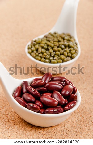 Different varieties of beans in ceramic spoons on a cork board: red kidney beans, mung beans
