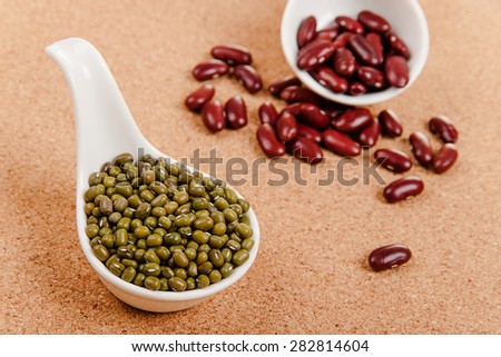 Different varieties of beans in ceramic spoons on a cork board: red kidney beans, mung beans