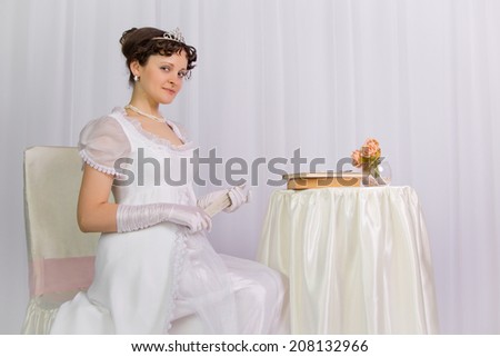 Young lady with a fan beside the table with a book and flowers, girl with hair in a ball gown, empire style