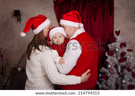 Dad, mom and baby in santa hat in anticipation of a happy Christmas. Family near Christmas tree, huddled together, the parents kiss
