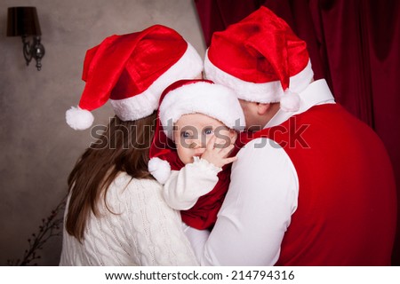 Dad, mom and baby in santa hat in anticipation of a happy Christmas. Family near Christmas tree, huddled together, the parents kiss