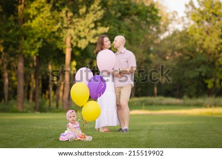Dad, mom and baby for a walk in the park. Baby with balloons in the foreground, Mom and Dad hugging in the background. Baby in focus, the parents are not in focus
