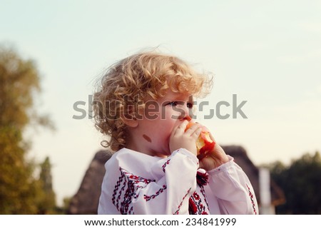Blonde curly toddler eats red apple in nature. Baby clothes in Ukrainian embroidery
