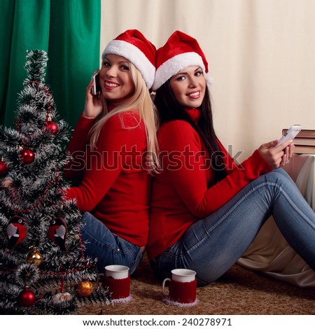 Two attractive young women friends near Christmas tree drinking tea and congratulate your friends and family by phone