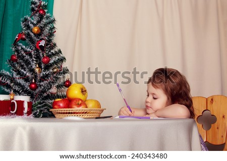 Little attractive girl writes a letter to Santa at the table next to the Christmas tree. New Year, Christmas