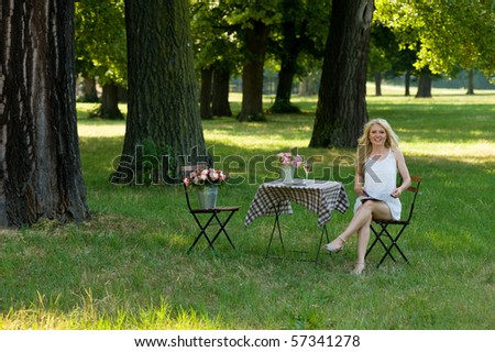 young blond in the park under trees, sitting relaxed on a folding chair beside a folding table with flowers and a wine glass on it