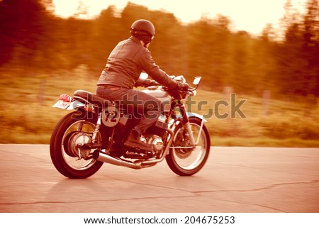 Young man riding a vintage motorcycle converted to a cafe racer. Fake license plate number. Short depth of field. Camera panning for motion blur.