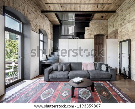 Interior in a loft style with brick walls, wooden ceiling and a parquet with a carpet on the floor. There is a gray sofa with pillows, round table, stair, windows, doors, white lockers, mirror.