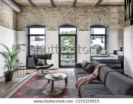 Interior in a loft style with brick walls, wooden ceiling and a parquet with carpet on the floor. There is a gray sofa with pillows and plaid, round table, stand, windows, green plant, stair, door.
