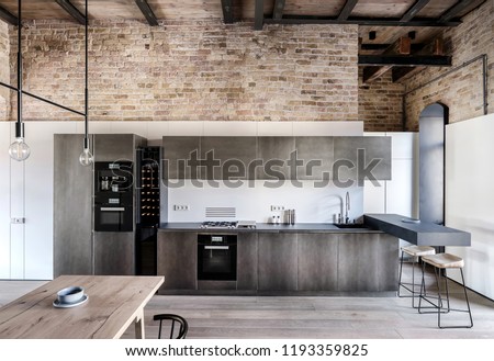 Kitchen in a loft style with brick walls, wooden ceiling and a parquet on the floor. There is wooden table, metal lockers, fridges, stove, oven, sink, dark tabletop, bar chairs, coffee machine.