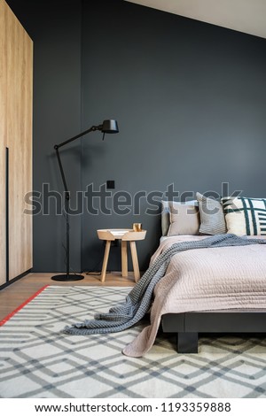 Stylish modern bedroom with dark and light walls and a parquet with a carpet on the floor. There is a bed with pillows and a coverlet and a plaid, black floor lamp, wooden wardrobe, small round table.