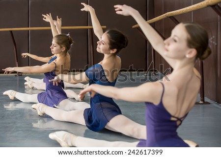 Young ballerinas on the lesson of classical choreography sitting in the splits gracefully raising his hands up near barre.
