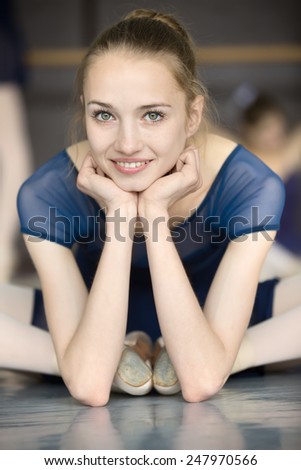 Young ballerina sitting on the floor with his face to the camera in the splits and dance class dancers practicing on the background. Close-up portrait