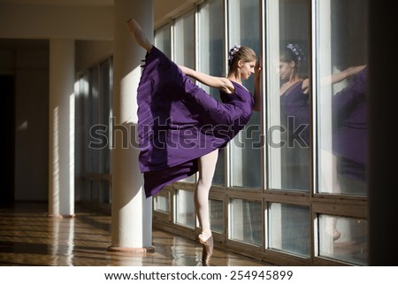 Graceful ballerina dancing in a purple dress leg lifted high, standing on pointe near a large window in the setting sun.