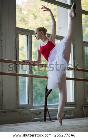 Young ballerina standing on one leg on your toes in pointe and doing stretching, lifting the leg up high. Model holds hands behind the barre.