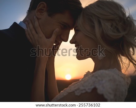 Close portrait silhouette in love wedding couple. Against the setting sun in the field. Lovers hug, girl tenderly holding her husband\'s face with both hands, smiling and looking into his eyes.