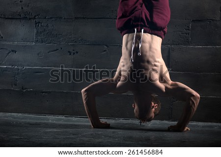 Athlete with naked torso doing push-ups on his hands while standing upside down near the concrete wall. Studio shots in the dark tone.