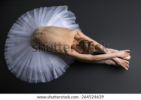 Young, graceful ballerina sitting on the floor on a black background in a studio in the slope face touching knees.