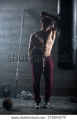 Young athlete with the ball in one hand stands near the punching bag. Snapshot in dark colors.
