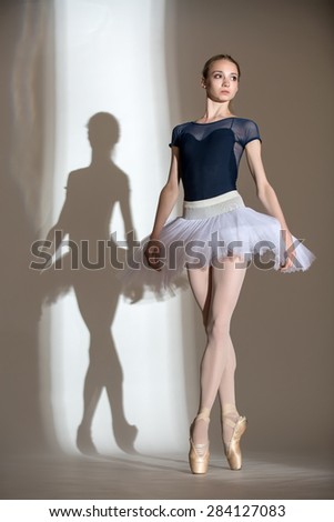Portrait in full growth graceful ballerina in a studio on a white background. Dancer in a blue bathing suit and white tutu. Against the background of her beautiful shadow.