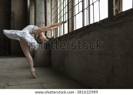 Young ballet dancer posing next to windows in an old industrial building. She is standing on pointes while she holds the grille during tilting back. Indoor. Horizontal.