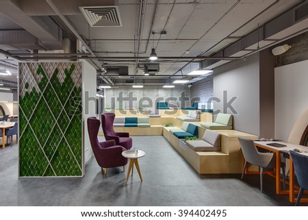 Interior in a loft style with a big zone with wooden benches and seats. Benches are decorated with grass, plants and bricks. On the right there is a gray table with orange legs and two chairs. In the