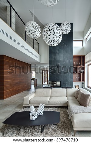 Hall in a modern style with light walls and big white round decorative lamps at the top. There is a beige sofa with pillows and plaid, dark table with three decorative vases, two black armchairs