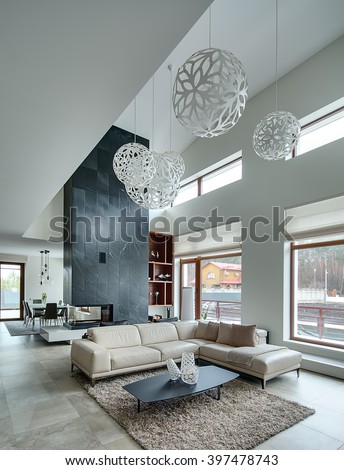 Hall in a modern style with light walls and big white round decorative lamps at the top. In the centre there is a beige sofa with pillows and plaid, dark table with three decorative vases, two black
