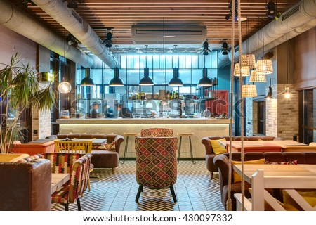Glowing interior in a loft style in a mexican restaurant with open kitchen on the background. In front of the kitchen there are wooden tables with multi-colored chairs and sofas. On the sofas there